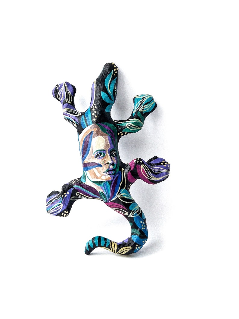 May include: A black paper mache lizard sculpture with a painted human face and colorful floral patterns. The lizard is in a curled up position with its tail curled around its body.