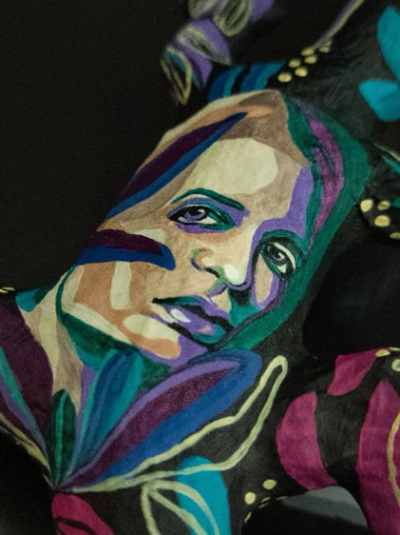May include: A close-up of a person's face painted with colorful abstract designs. The face is painted with shades of blue, green, purple, and yellow. The person's eyes are closed and their lips are painted a light purple.