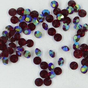 May include: A scattering of small, round, red rhinestones with iridescent blue and purple facets. The rhinestones are scattered on a white surface.
