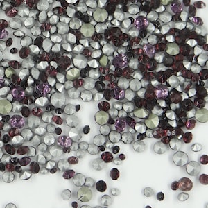 Op de afbeelding: Een mix van zilveren, paarse en bordeauxrode strass steentjes. De strass steentjes zijn verspreid over een wit oppervlak.