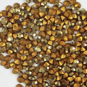 Peut inclure: Une grande quantité de strass dorés, également appelés pierres précieuses en verre, sont éparpillés sur une surface blanche. Les strass ont un design facetté et une finition métallique.