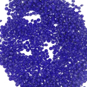 Peut inclure: Un tas de strass bleu foncé à facettes. Les petits morceaux en forme de gemmes sont dispersés sur une surface blanche, créant un effet scintillant. Ces fournitures artisanales sont idéales pour divers projets de bricolage.