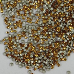 Peut inclure: Gros plan sur une dispersion de petits strass à facettes. Les strass sont un mélange de couleurs argentées et dorées, créant un effet scintillant. Les pierres précieuses sont dispersées sur une surface blanche, avec une variété de formes et de tailles.