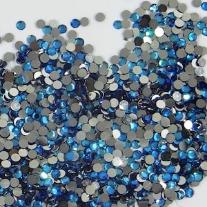 May include: A large quantity of small, round, blue and silver rhinestones. The rhinestones are scattered on a white surface.