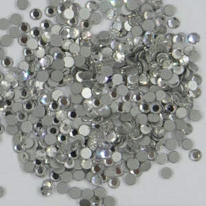 May include: A large pile of silver-coloured rhinestones, also known as crystals or gems, are scattered on a white surface. The rhinestones are round and have a faceted design.