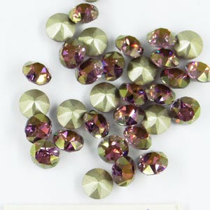 Pode incluir: Uma variedade de strass iridescentes em várias formas, incluindo redondas e ovais. Os strass têm uma base prateada metálica e uma mistura brilhante de tons roxos, rosa e verdes. As pedras preciosas estão espalhadas sobre uma superfície branca.