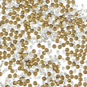 May include: A scattering of small, faceted rhinestones in gold and clear colors. The rhinestones are round with flat backs and are scattered across a white background. The gold rhinestones have a metallic sheen.