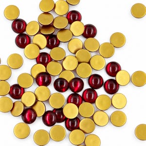 Peut inclure: Une collection de cabochons circulaires comprenant un mélange de disques dorés mats et de dômes en verre rouge profond translucide. Les pièces sont dispersées, montrant un contraste entre la finition métallique et les surfaces rouges lisses.