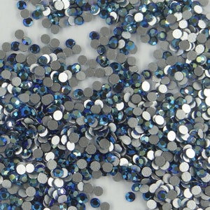 Puede incluir: Una gran cantidad de pequeñas piedras de cristal redondas de color plateado con centros azules iridiscentes. Las piedras de cristal están dispersas sobre una superficie blanca.