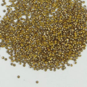 Könnte beinhalten: Nahaufnahme eines Haufens kleiner, facettierter, goldfarbener Strasssteine. Die Strasssteine haben einen metallischen Glanz und sind auf einer weißen Oberfläche verstreut. Die Edelsteine haben unterschiedliche Größen und Formen und erzeugen ein strukturiertes Aussehen.