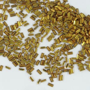 May include: A large quantity of gold-colored rectangular rhinestones. The rhinestones are scattered on a white surface.