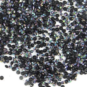 Puede incluir: Primer plano de un montón de pedrería negra iridiscente. Las piedras redondas y facetadas capturan la luz, creando un efecto brillante. Las gemas están dispersas sobre una superficie blanca.