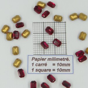 Könnte beinhalten: Nahaufnahme von roten und goldenen Glasperlen. Die Perlen sind in einem zufälligen Muster auf einer weißen Oberfläche angeordnet. Im Hintergrund befindet sich ein Lineal mit Millimetermarkierungen. Der Text "Papier millimétré. 1 carré = 10mm 1 square = 10mm" ist auf einem weißen Etikett gedruckt.
