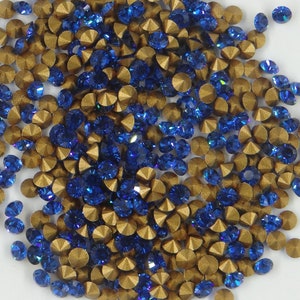 Peut inclure: Gros plan sur une collection de petits strass étincelants. Les strass sont un mélange de bleu profond et d'or, dispersés sur une surface blanche. Les strass bleus sont facettés, captant la lumière, tandis que les dorés ont une finition métallique.