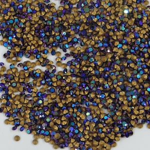 Peut inclure: Une grande quantité de strass dorés et violets irisés. Les strass sont éparpillés sur une surface blanche.