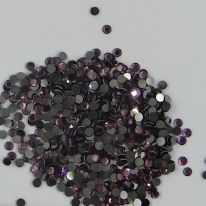 Peut inclure: Un grand tas de petits strass ronds, irisés, violets et noirs. Les strass sont éparpillés sur une surface blanche.