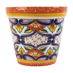 Op de afbeelding: Een kleurrijke keramische plantenpot met een witte achtergrond en een geometrisch patroon van blauwe, gele, oranje en paarse bloemen. De pot heeft een gegolfde rand en een gele band rond de bovenkant.