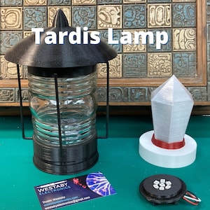 Könnte beinhalten: Eine schwarze Tardis-Lampe mit einer klaren Glaskugel und einem weißen Sockel. Die Lampe steht neben einer weißen und roten Tardis-förmigen Lampe mit silbernem Finish. Die Lampen stehen auf einer grünen Oberfläche mit einer Visitenkarte für Westaby Electronics.