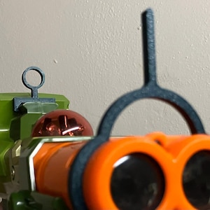 以下が含まれることがあります： 黒色のスコープと赤と金のバレルが付いた緑とオレンジの玩具銃。銃の上部に黒い照準器、側面に黒いリングが付いている。