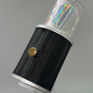 Puede incluir: Objeto cilíndrico negro y plateado con una parte superior transparente y redondeada. La parte superior contiene un diseño holográfico con colores del arcoíris. Un botón dorado está en el lateral. Probablemente sea un artículo decorativo o un accesorio único.