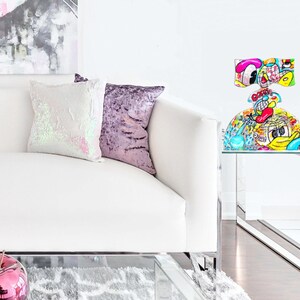 May include: A white couch with two pillows, one with iridescent sequins and the other with a purple velvet fabric. A glass end table with a colorful, cartoon-style glass sculpture on top. The sculpture has a round base and a top that resembles a bow. The sculpture is decorated with bright colors and cartoon characters.