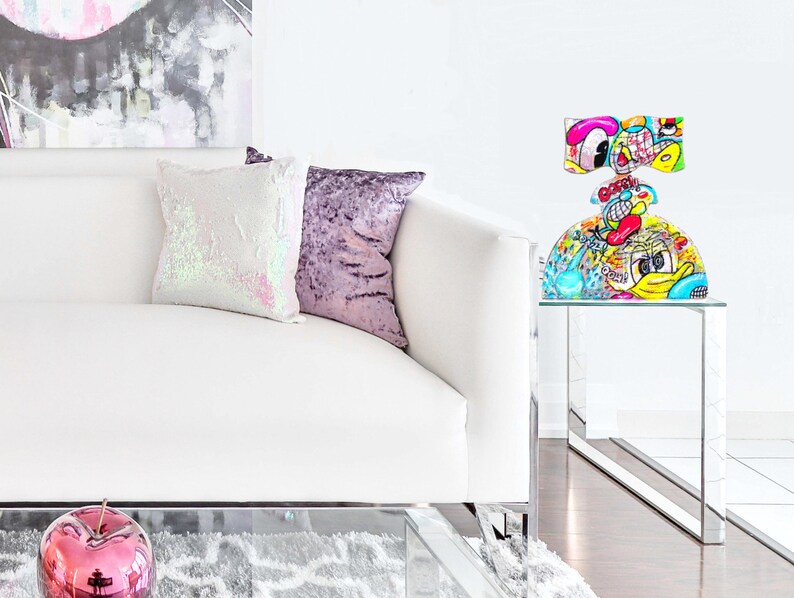 May include: A white couch with two pillows, one with iridescent sequins and the other with a purple velvet fabric. A glass end table with a colorful, cartoon-style glass sculpture on top. The sculpture has a round base and a top that resembles a bow. The sculpture is decorated with bright colors and cartoon characters.