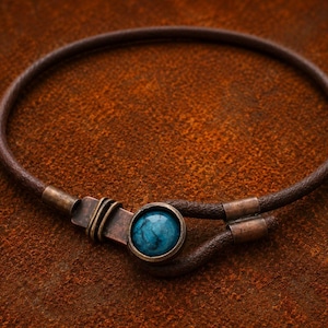 Op de afbeelding: Een ketting van donkerbruin leer met een ronde sluiting van koperkleurig metaal. De sluiting is voorzien van een enkele ronde, gepolijste turkooiskleurige steen in een zetting, met decoratieve draadwikkelingen op de metalen verbinding.