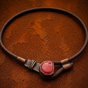 Op de afbeelding: Een ketting van donkerbruin leren koord met een centrale gepolijste rode steen in een verweerde koperen zetting. De ketting bevat koperen accenten met een gewikkeld draaddetail en een haaksluiting aan de zijkant.