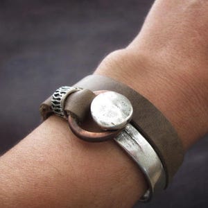 May include: A brown leather bracelet with a silver and copper clasp. The bracelet is wrapped around a person's wrist.