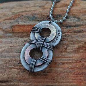 May include: A silver and copper necklace with a double ring pendant. The rings are intertwined with wire and have a small copper dot in the center. The necklace is on a wooden surface.