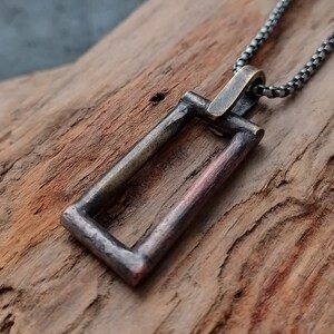 May include: A silver chain necklace with a rectangular pendant. The pendant is made of metal with a dark, aged finish and a copper-colored interior.