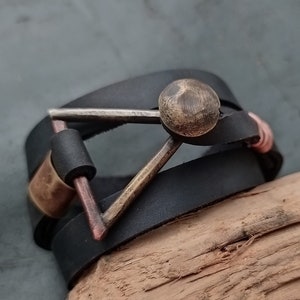 Puede incluir: Un brazalete de cuero negro con un cierre triangular de metal y un acento redondo de metal. El brazalete está envuelto alrededor de un trozo de madera a la deriva.