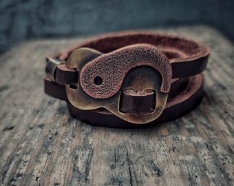May include: A brown leather bracelet with a brass buckle featuring a textured, circular design. The bracelet is wrapped twice around the wrist.
