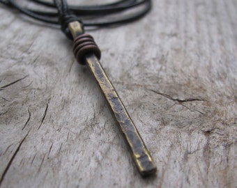 May include: A long, thin, hammered brass pendant hangs from a black leather cord. The pendant has a rough, textured surface and is a simple, minimalist design.