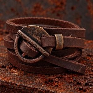 May include: A dark brown leather wrap bracelet featuring a circular bronze-toned metal clasp. The clasp has a textured, weathered finish with small etched markings along the outer ring. The leather strap is flat and wraps multiple times around.