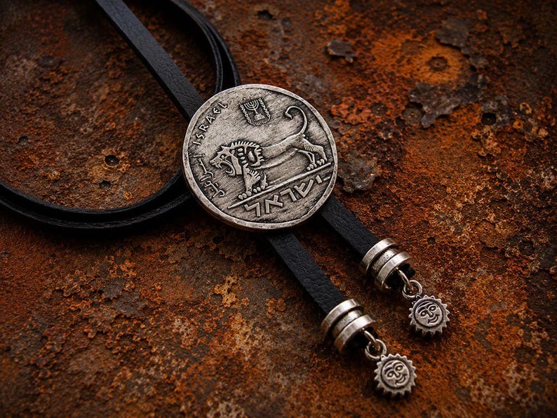 May include: A bolo tie featuring a circular silver-toned coin pendant embossed with a lion, the word ISRAEL, and a menorah symbol. The pendant is attached to a black leather cord finished with two silver-toned metal tips, each featuring a sun charm.