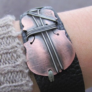 May include: A black leather bracelet with a copper-colored violin cutout. The violin is decorated with silver wire.