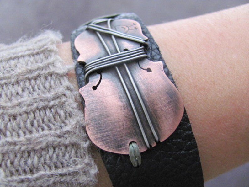 May include: A black leather bracelet with a copper-colored violin cutout. The violin is decorated with silver wire.