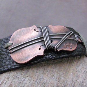 May include: A black leather bracelet with a copper and silver violin-shaped charm. The charm is decorated with wire and has a small bird design.