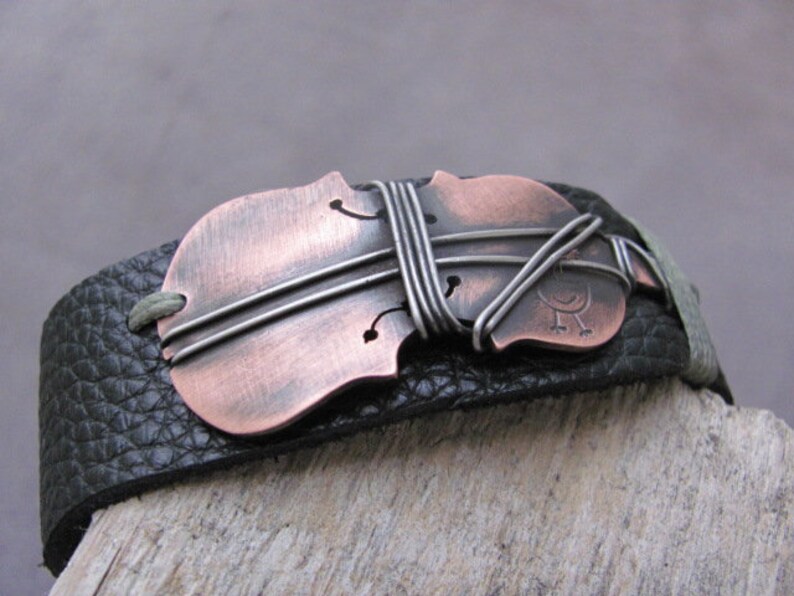 May include: A black leather bracelet with a copper and silver violin-shaped charm. The charm is decorated with wire and has a small bird design.