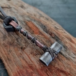 May include: A silver and copper wire-wrapped hammer pendant necklace. The hammer is made of metal and has a rounded head. The pendant is attached to a brown leather cord.
