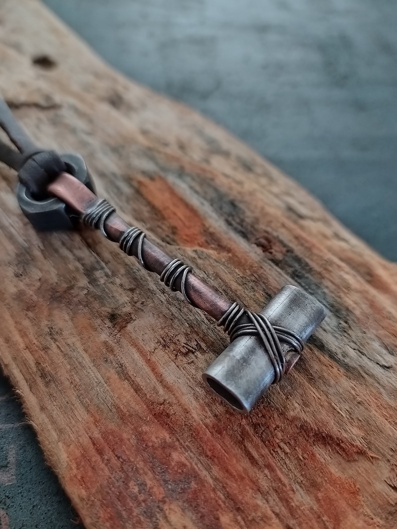 May include: A silver and copper wire-wrapped hammer pendant necklace. The hammer is made of metal and has a rounded head. The pendant is attached to a brown leather cord.