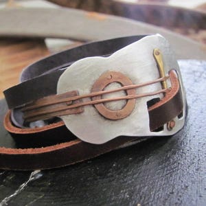 May include: A leather bracelet with a silver guitar-shaped charm. The charm has a copper inlay and a brass accent.