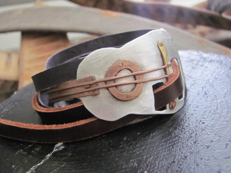 May include: A leather bracelet with a silver guitar-shaped charm. The charm has a copper inlay and a brass accent.