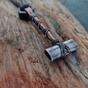 May include: A silver and copper necklace with a small hammer pendant. The hammer is made of metal and has a textured surface. The necklace is on a wooden background.