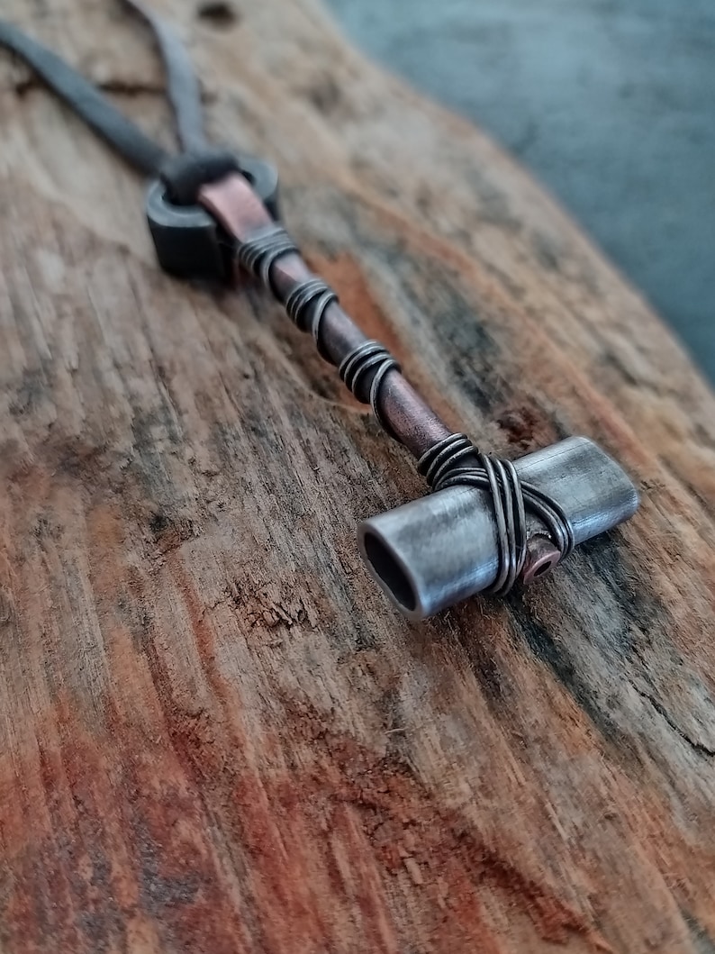 May include: A silver and copper necklace with a small hammer pendant. The hammer is made of metal and has a textured surface. The necklace is on a wooden background.
