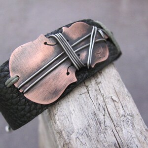 May include: A black leather bracelet with a copper-colored violin cutout. The violin is accented with silver wire.