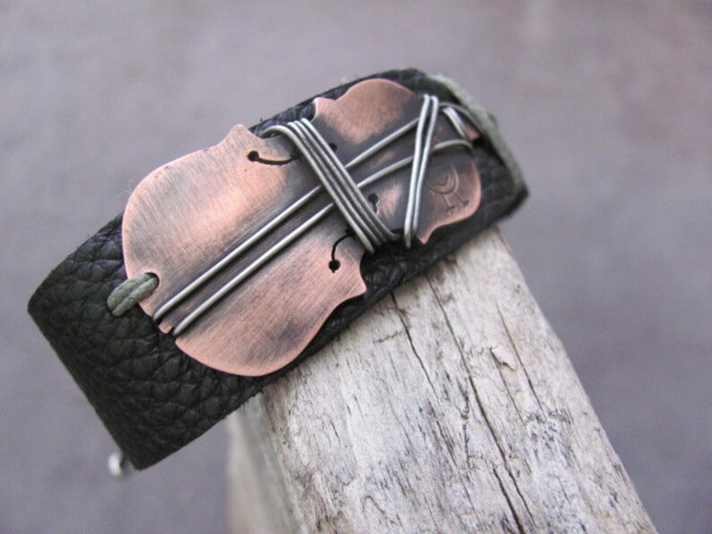 May include: A black leather bracelet with a copper-colored violin cutout. The violin is accented with silver wire.