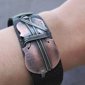 May include: A black leather bracelet with a copper-colored violin-shaped charm. The charm is decorated with silver wire and has a distressed finish.
