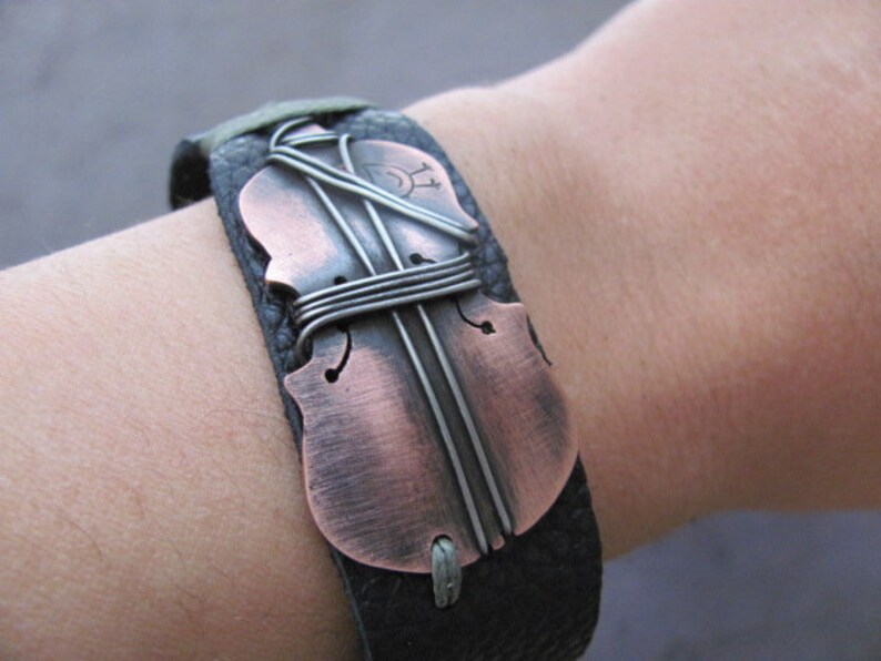 May include: A black leather bracelet with a copper-colored violin-shaped charm. The charm is decorated with silver wire and has a distressed finish.